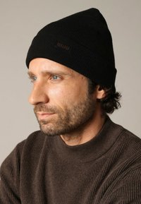 Zwart gebreide muts met een klein bruin label, gedragen door een persoon; gestructureerd materiaal, aansluitend ontwerp. Bruine geribbelde trui zichtbaar eronder.