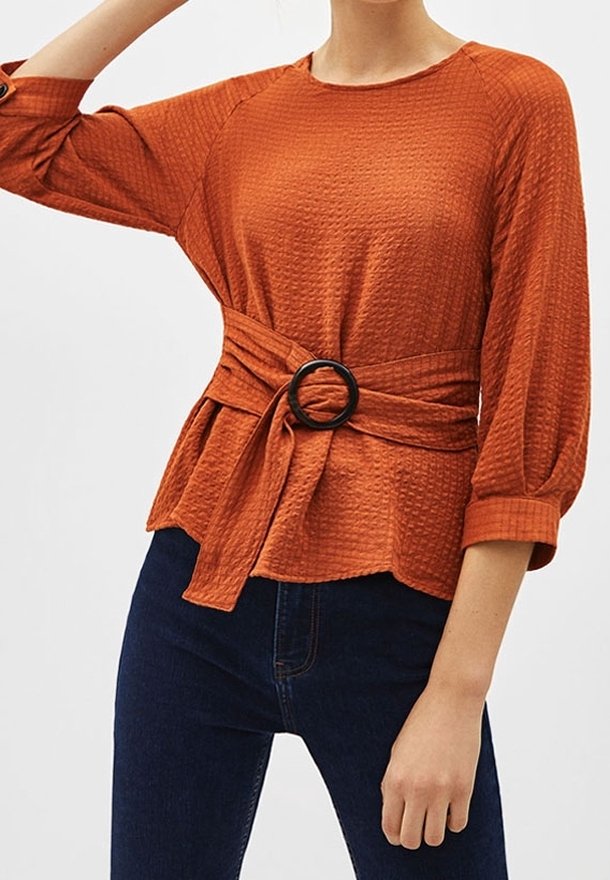Kvinde iført en tekstureret rust-orange langærmet bluse med sort cirkulær bæltespænde, parret med mørkeblå jeans, mod en ensfarvet baggrund.