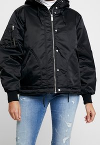 Giubbotto bomber in nylon nero con cappuccio, chiusura a zip sul davanti, due tasche laterali e una tasca con zip sulla manica. Polsini a costine e orlo regolabile con cordino.