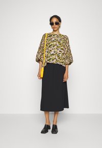 Blusa larga de padrão floral com mangas bufantes em verdes e rosas, combinada com uma saia maxi preta e tamancos pretos. Bolsa transversal amarela.