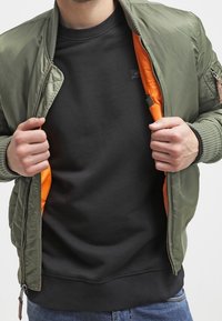 Chaqueta bomber de color verde oliva con puños y cintura acanalados, desabrochada para revelar un forro naranja. Llevada sobre una sudadera negra.