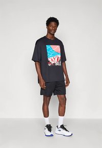 Άνδρας που στέκεται, φοράει μαύρο υπερμεγέθες μπλουζάκι με θέμα το μπάσκετ, μαύρο σορτς, λευκές κάλτσες και λευκά αθλητικά παπούτσια με μπλε σόλες.