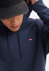 Marineblauwe hoodie met een capuchon en trekkoord, met een klein rood Levi's logo. Donker getextureerde pet achterstevoren gedragen, met een vlakke rand.