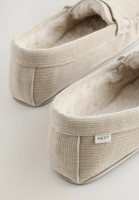Beige corduroy slippers met zachte nep bontvoering, een platte zool en een klein label met "NEXT" aan de zijkant. Rond, instapontwerp.