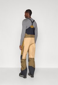 Férfi, aki többszínű síbabákat visel felfüggesztőkkel és szürke hosszú ujjú pólóval, háttal áll, egyszínű háttér előtt.