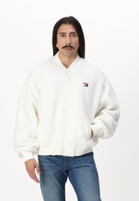 Chaqueta bomber blanca hecha de suave forro polar texturizado, con cierre de cremallera, puños acanalados y un pequeño parche con logo en el pecho.