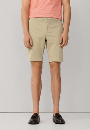 Mann trägt beige knielange Shorts, pfirsichfarbenes T-Shirt und dunkelbraune Tassel-Loafer, steht vor einem schlichten hellen Hintergrund.