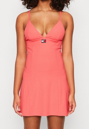Woman wearing a coral pink ribbed mini dress with thin straps and a "Tommy Jeans" logo patch on the chest.