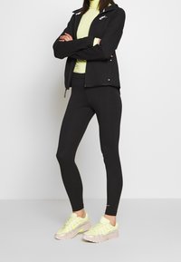 Person stands with arms crossed wearing black Nike jacket, black leggings, light green turtleneck, and beige and neon yellow sneakers.