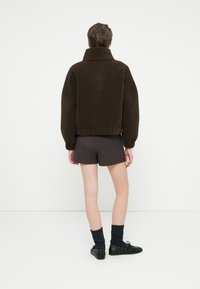 Jachetă din fleece maro cu guler înalt și croială supradimensionată, purtată cu pantaloni scurți negri și pantofi negri. Vedere din spate, evidențiind materialul texturat.