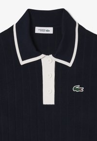 Polo en maille bleu marine avec un col et une patte de boutonnage blancs. Il dispose de trois boutons et d'un petit logo crocodile vert sur la gauche.