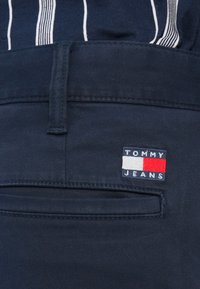 Κοντινή εικόνα στην τσέπη σκούρου μπλε παντελονιού που δείχνει το λογότυπο Tommy Jeans και μέρος από ριγέ σκούρο μπλε και λευκό πουκάμισο.