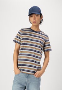 T-shirt rayé en beige, bleu et marron avec des manches courtes, un col rond et un écusson rouge. Assorti avec un jean bleu clair et une casquette marine.
