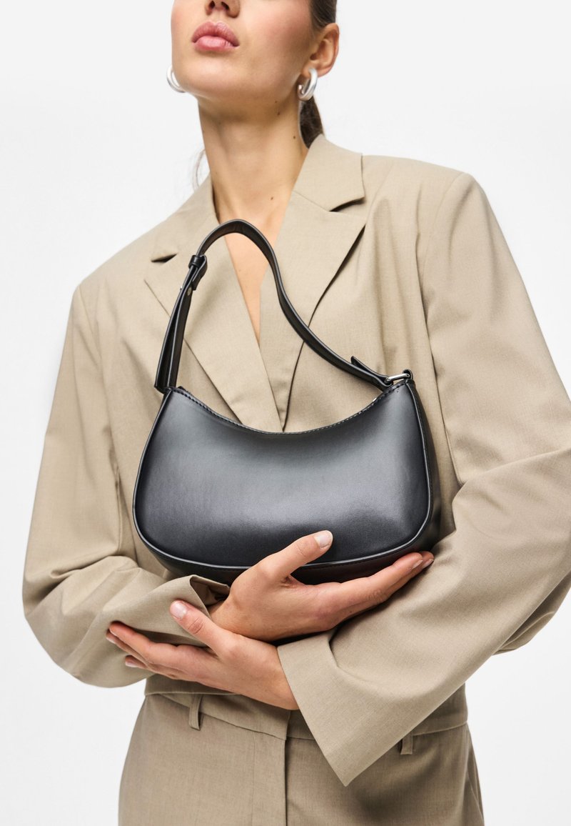 Femme en blazer beige tenant un petit sac à bandoulière en cuir noir avec une sangle courte sur fond blanc.