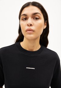 Schwarzes Baumwoll-T-Shirt mit Rundhalskragen und kurzen Ärmeln, mit weißem "ARMEDANGELS"-Logo auf der Brust. Glatte Textur.
