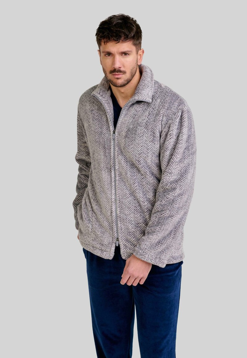 Chaqueta gris de espiga con cremallera y cuello alto, con bolsillos laterales, confeccionada en una tela suave. Combinada con pantalones suaves azul marino.