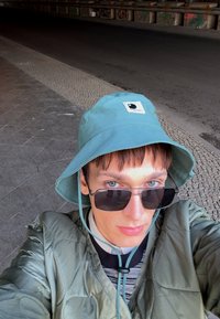 Teal bucket hat med et logo patch, olivengrøn polstret jakke, sorte solbriller og stribet skjorte, sat mod en tekstureret urban baggrund.