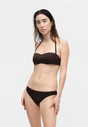 Vrouw met nat haar die een donkerbruine strapless bikini top met halterbandjes en bijpassende bikinibroek draagt tegen een effen lichte achtergrond.