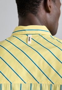 Žluté pruhované tričko s modrými pruhy, které má límec a všitou etiketu na zadní straně. Tkanina vypadá hladce a lehce.
