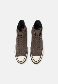 Brune høyhælte sneakers laget av teksturert stoff, med hvite gummitupper, svarte såler og oransje Gore-Tex-merke på siden.