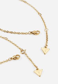 Collier en chaîne dorée avec plusieurs maillons, fermoirs en forme de homard, et un pendentif triangulaire portant un logo gravé.