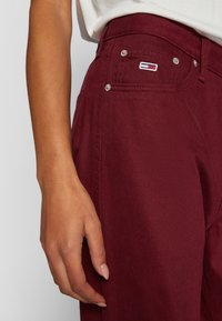 Pantalones de mezclilla granate con herrajes plateados, mostrando un bolsillo frontal y un parche con el logo en la cinturilla. Detalles de textura y costuras visibles.