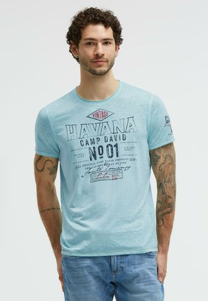 Mann mit lockigem Haar, der ein hellblaues "Havana No 01" T-Shirt und Jeans trägt, zeigt Tattoos an beiden Armen und steht vor einem einfarbigen Hintergrund.