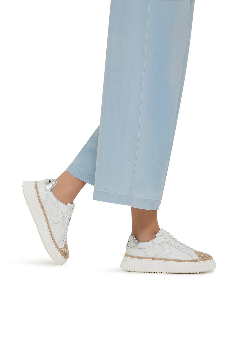 Sneakers bianchi con una punta strutturata beige e accenti argentati, caratterizzati da una suola spessa e rigata e lacci, abbinati a pantaloni a gamba larga blu chiaro.