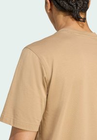 Tan katoenen t-shirt met korte mouwen, platte naden en een ronde hals. De achterkant toont een glad, onbewerkt oppervlak.