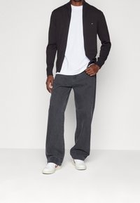 Czarna kurtka rozpinana na suwak na białej koszulce, w połączeniu z luźnymi ciemnoszarymi dżinsami i białymi trampkami z czerwonymi akcentami.