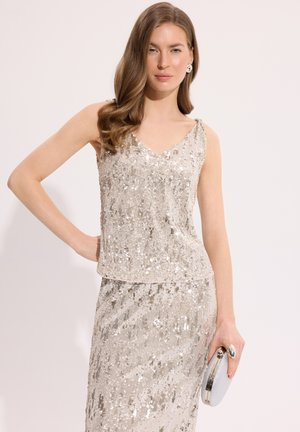 Kobieta z długimi falowanymi włosami, ubrana w srebrny, cekinowy, bezrękawnik oraz pasującą spódnicę, trzymająca w prawej ręce małą srebrną kopertówkę.