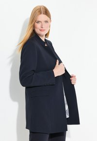 Blazer navy con colletto alto, chiusura frontale a bottoni e tasche, realizzato in tessuto liscio, indossato aperto su una maglietta a fantasia.