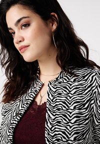 Zwart-wit zebraprint jas met een gestructureerde stof, zilveren drukknoopjes, gedragen over een bordeauxrode kanten top en fijne gouden sieraden.