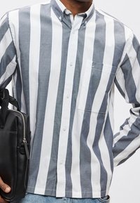 Homme portant une chemise à rayures verticales grises et blanches avec boutons, tenant un sac en cuir noir avec fermeture éclair.