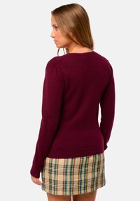 Weinroter Pullover mit langen Ärmeln und rundem Halsausschnitt, kombiniert mit einem beigen Karo-Rock, der rote und grüne Akzente hat. Sanfte Textur.
