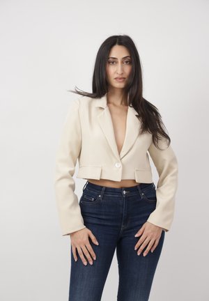 Frau mit langem dunklem Haar, die einen kurzen beigen Blazer und dunkelblaue hoch taillierte Jeans trägt und vor einem schlichten weißen Hintergrund steht.