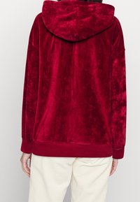 Felpa in velluto rosso con finitura testurizzata, grande cappuccio e polsini a coste. Il capo ha una vestibilità ampia ed è abbinato a pantaloni in velluto a coste color crema.