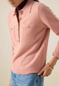 Pull en tricot rose avec un col, présentant un motif de côtes verticales, deux poches poitrine avec des boutons dorés, et des poignets et un ourlet côtelés.