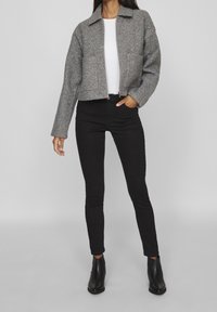 Szara przycięta kurtka z teksturowanej tkaniny, z przodu zamek błyskawiczny i kołnierz, w połączeniu z czarnymi obcisłymi jeansami i czarnymi botkami na kostkę.