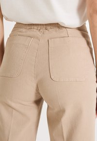 Pantalones beige de cintura alta con un acabado texturizado, que presentan dos bolsillos traseros y una cintura fruncida para un ajuste relajado.