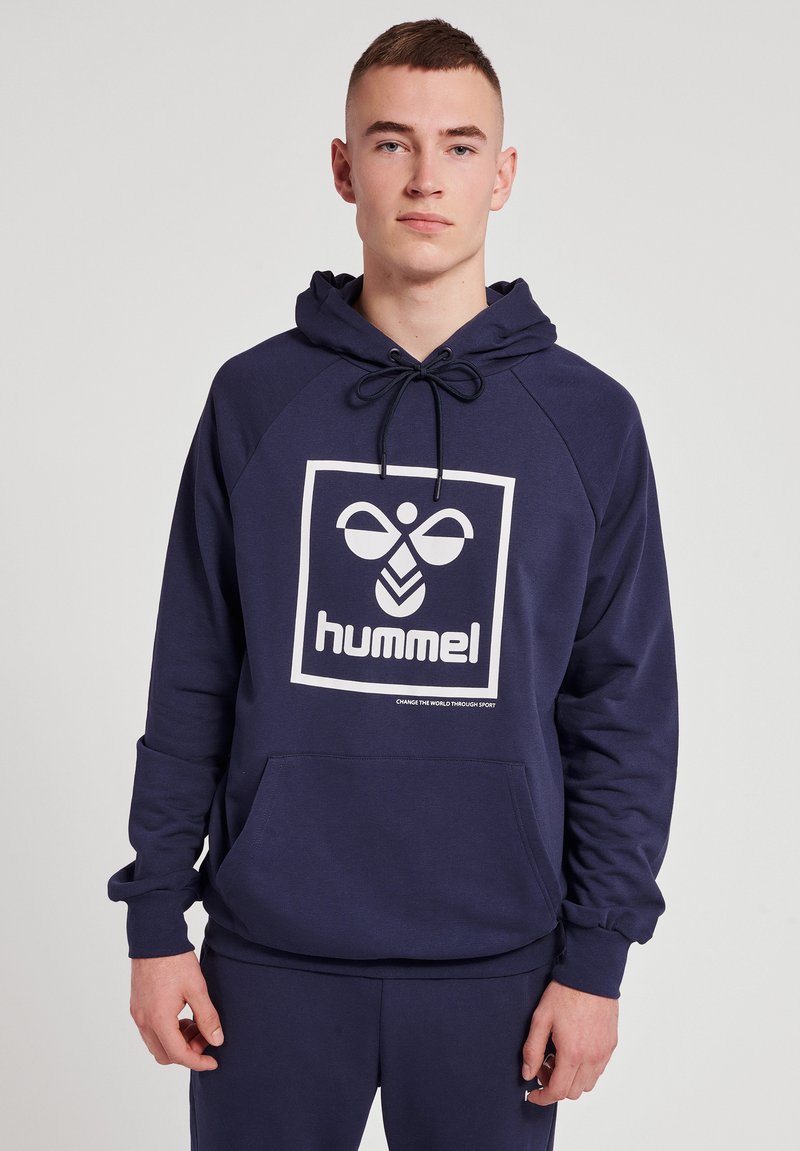 Młody mężczyzna w granatowej bluzie z kapturem z białym logo Hummel i kieszenią kangurką, stojący na jednolitym jasnoszarym tle.