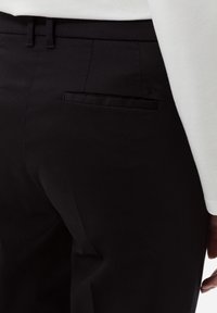 Pantaloni neri caratterizzati da un design elegante, una texture liscia e una tasca posteriore con un logo discreto. La vita è dotata di passanti per cintura.