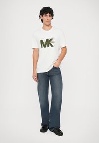 Jauns vīrietis, tērpies baltā Michael Kors logotipa T-kreklā, zilos džinsos un melnās kedās, stāv ar vienu roku kabatā uz vienkārša fona.