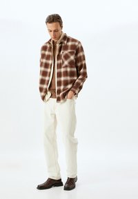 Camicia di flanella a quadri marrone con accenti bianchi, sovrapposta a una camicia marrone, abbinata a pantaloni beige chiaro e stivaletti marroni.