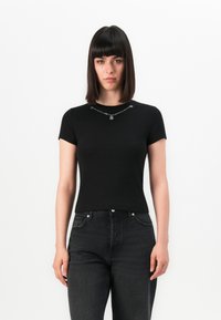 Woman with straight dark hair wearing a black short-sleeve top, silver chain necklace, and high-waisted dark jeans against a white background.