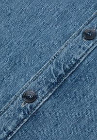 Zbliżenie na niebieską tkaninę dżinsową z dwiema ciemnymi guzikami na przeszytej klapie, ukazujące teksturę oraz szczegóły otworów na guziki.