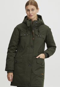 Parka verte de longueur moyenne en tissu texturé. Elle possède une capuche, des fermetures éclair et à bouton, deux poches poitrine et un ourlet ajustable avec cordon.
