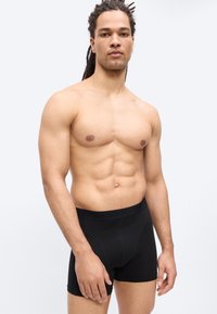 Homme musclé aux dreadlocks portant un caleçon noir, debout devant un fond blanc uni, regardant droit devant.