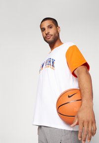 Fehér, rövid ujjú póló narancssárga részletekkel és "NEW YORK" felirattal, szürke rövidnadrággal párosítva. Narancssárga kosárlabdát tart, rajta fekete Nike logóval.