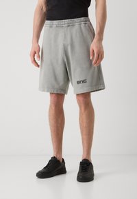 Pantalones cortos de algodón gris con cinturilla elástica, bolsillos laterales y un logotipo negro en la pierna izquierda. Combinados con zapatillas negras.
